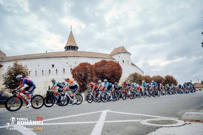 Cicliști în cursa de la Brașov la Ploiești, cu cetate veche în fundal.