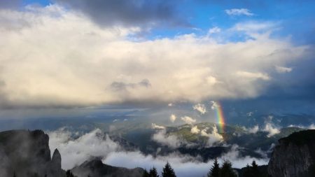 Peisaj montan cu nori, cer senin și curcubeu în fundal.