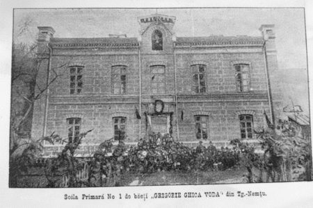 Clădire istorică a școlii primare din Târgu-Neamț, înconjurată de flori și copaci vechi.