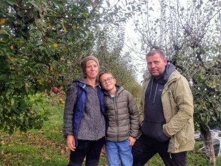 Ogorul soarelui din Sagna. Istoria legumelor faimoase ale soților Bordea! Trei persoane în livada de mere, înconjurate de pomi fructiferi și frunze verzi.
